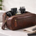 Brown leather toiletry bag on a bathroom countertop with grooming essentials like razor, cologne, and comb arranged around it